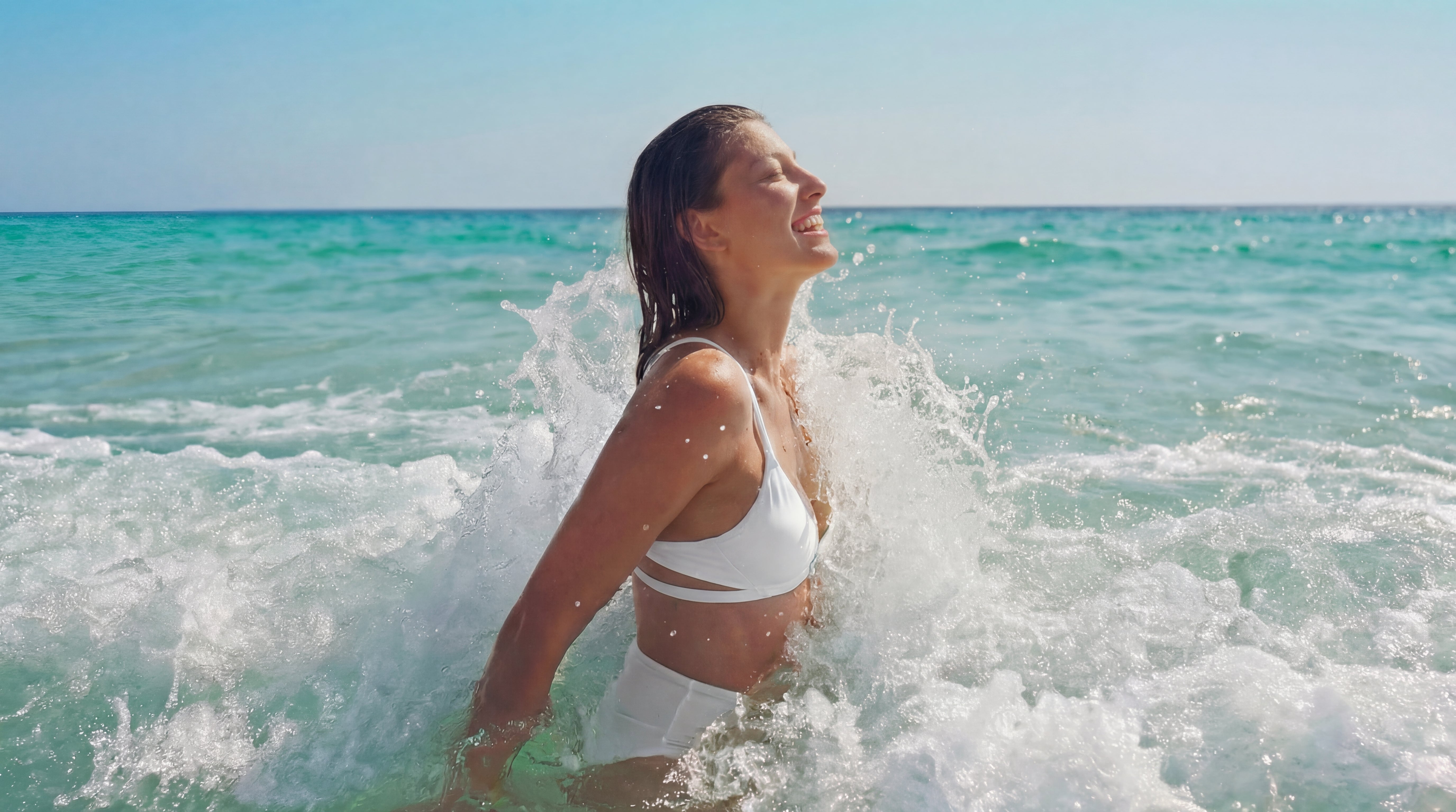 Woman in a white bikini standing in the ocean with waves crashing around her.